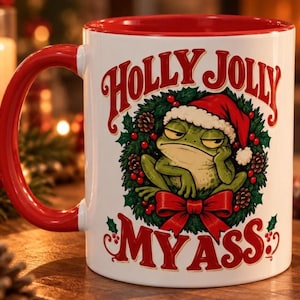 Könnte beinhalten: Ein weißer Keramikbecher mit rotem Henkel und rotem Innenraum. Auf der Vorderseite ist ein grüner Frosch mit Weihnachtsmütze in einem Weihnachtskranz mit roter Schleife abgebildet. Der Text lautet HOLLY JOLLY MY ASS in roter, dekorativer Schrift.