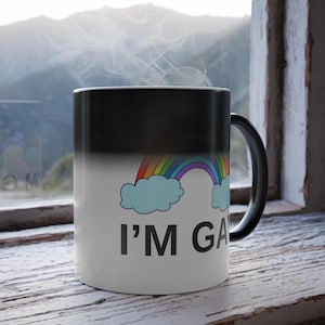 Puede incluir: Una taza que cambia de color, negra en la parte superior y blanca en la inferior, con un arcoíris y nubes. La taza muestra el texto "I'M GAY". Sale vapor de la taza, lo que sugiere una bebida caliente.