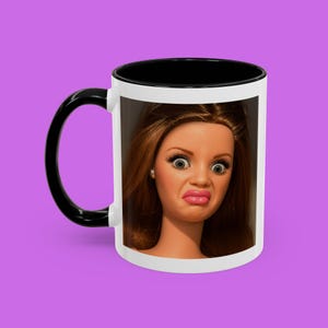 May include: White ceramic mug with a black handle and interior. The mug features a close-up of a doll's face with a surprised expression. The doll has brown hair, large eyes, and pink lips. The mug sits on a purple background.