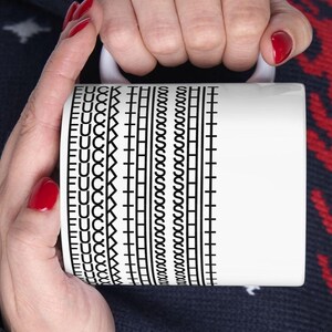 May include: White ceramic mug with a black graphic pattern and the word "FUCK" repeated vertically. The mug is held by a person with red nail polish.