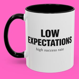 May include: White ceramic mug with a black interior and handle. The mug features the text "LOW EXPECTATIONS" in bold black letters, with "high success rate" below it. The mug is set against a pink background.