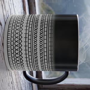 May include: A white and black patterned ceramic mug with a black handle. The mug's design includes repeating patterns of U-shaped, zigzag, and square shapes. Steam rises from the dark side of the mug, suggesting a hot beverage.