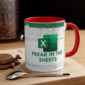 May include: A white ceramic mug with a red interior and handle. The mug features a green and white Excel spreadsheet design with the text "FREAK IN THE SHEETS" in green. The mug is on a wooden surface with a spoon and a macaron.
