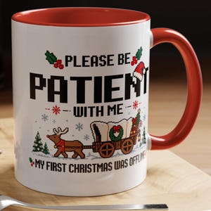 Könnte beinhalten: Ein weißer Keramikbecher mit rotem Innenraum und Henkel, verziert mit Pixel-Art-Grafiken eines Rentiers, das einen Planwagen zieht, sowie Stechpalmen und einer Weihnachtsmütze. Der Text lautet: PLEASE BE PATIENT WITH ME, MY FIRST CHRISTMAS WAS OFFLINE.
