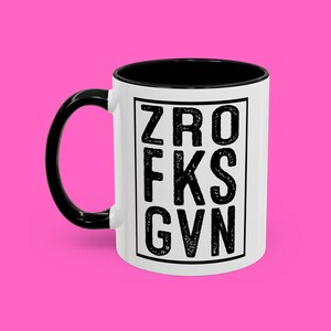 Op de afbeelding: Witte keramische koffiemok met een zwarte handgreep en een zwarte rand. De mok heeft een zwarte afbeelding van de letters "ZRO FKS GVN" die op de voorkant is gedrukt.