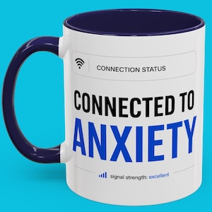May include: A white ceramic mug with a dark blue interior and handle. The front features a graphic resembling a digital interface with the text CONNECTION STATUS, CONNECTED TO ANXIETY, and signal strength: excellent with a signal icon.