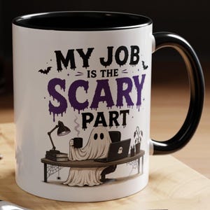 Op de afbeelding: Een witte keramische mok met een zwarte binnenkant en handvat, voorzien van een afbeelding van een geest aan een bureau met een laptop en een lamp. De tekst luidt "MY JOB IS THE SCARY PART", omringd door vleermuizen en spinnenwebben.
