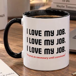 Puede incluir: Una taza de cerámica blanca con asa e interior negros, que presenta el texto negro "I LOVE MY JOB." repetido tres veces, seguido del texto rojo "Repeat as necessary until convinced." La taza está llena de una bebida líquida de color marrón oscuro.