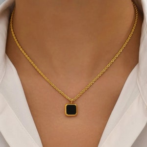 Peut inclure: Un collier doré avec un pendentif carré noir. Le pendentif, encadré d'or, est suspendu à une chaîne délicate. Le collier est présenté sur une chemise blanche.