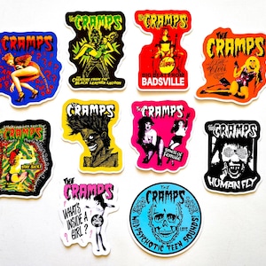 May include: A selection of vibrant stickers showcasing The Cramps band imagery. Designs include a skull, a creature, and other band-related graphics. The stickers are in various shapes and sizes, with bold colours and text.