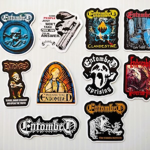May include: A selection of Entombed band stickers. Designs include a skull and crossbones, a revolver, and album art. The stickers are in various colours, including blue, black, red, and yellow. The stickers have a white border.