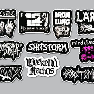 May include: A collection of eleven black and white vinyl stickers featuring various band logos and graphic illustrations. The designs include text such as ACxDC, XBRAINIAX, IRON LUNG, LARM, HUMMINGBIRD OF DEATH, SHITSTORM, MIND OF ASIAN, GANX, WEEKEND NACHOS, and GODSTOMPER.