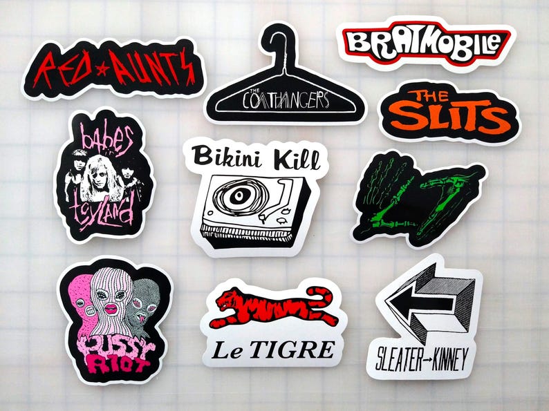 a collection of nine stickers, each with a unique design and text. The stickers are arranged in a grid-like pattern, with some overlapping each other. The stickers feature various artistic styles and themes, including a red and white logo, a black and white image of a tiger, a pink and black image of a woman, and a black and white image of a man. The stickers also include the words "Red Aunts", "The Slits", "Bikini Kill", "Le Tigre", and "Sleater-Kinney".