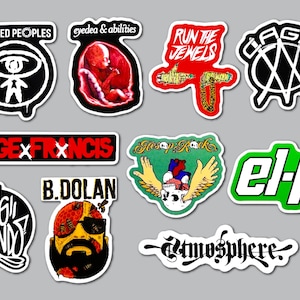 May include: A collection of ten vinyl stickers featuring various hip-hop artist logos and graphic designs. The stickers include text such as Dilated Peoples, Eyedea & Abilities, Run The Jewels, Cage, Sage Francis, Aesop Rock, El-P, Living Legends, B. Dolan, and Atmosphere.