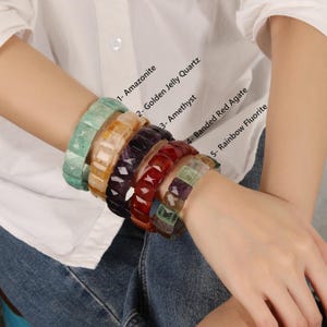 Könnte beinhalten: Fünf Edelsteinarmbänder in verschiedenen Farben: Grün, Gelb, Lila, Rot und Grün. Jedes Armband besteht aus facettierten rechteckigen Steinen. Die Abbildung zeigt die Namen der Edelsteine: Amazonit, Golden Jelly Quarz, Amethyst, Gebänderter Roter Achat und Regenbogen-Fluorit.