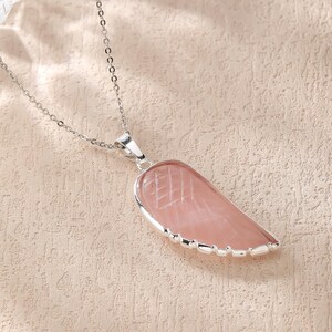 May include: A silver chain necklace with a pink, wing-shaped pendant. The pendant has a silver border and etched lines. The necklace is displayed on a textured, light-colored surface.
