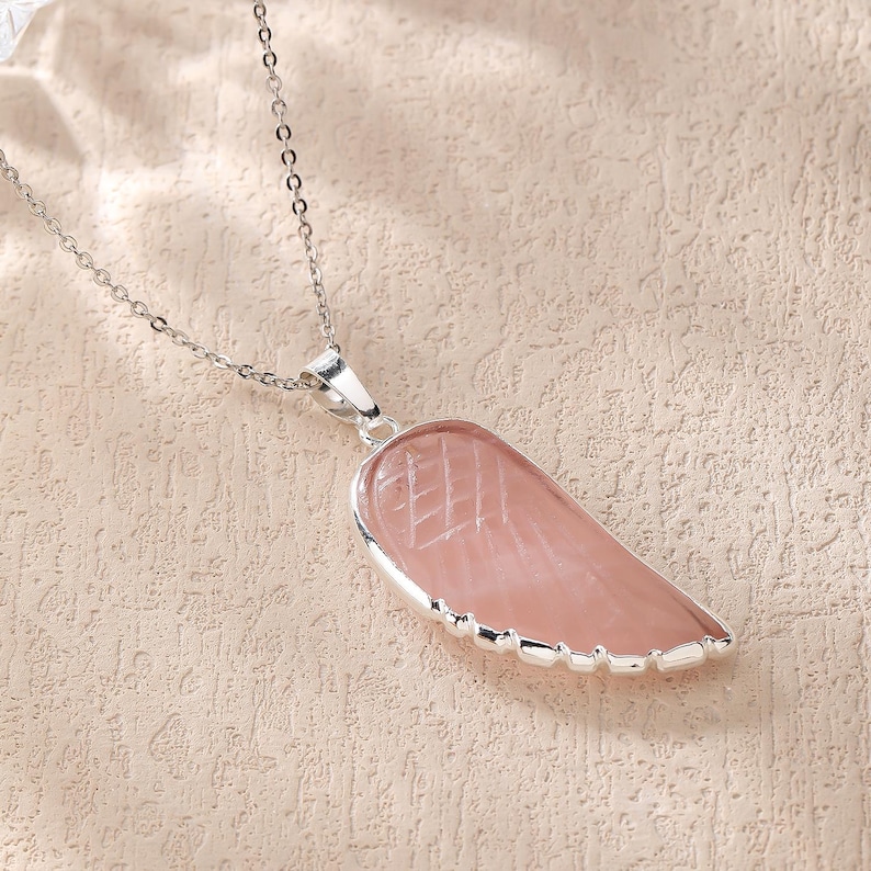 May include: A silver chain necklace with a pink, wing-shaped pendant. The pendant has a silver border and etched lines. The necklace is displayed on a textured, light-colored surface.