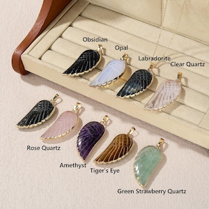 May include: A collection of gemstone wing pendants in various colors, including black obsidian, white opal, and green strawberry quartz. Each pendant has a gold-colored frame and is labeled with its gemstone type.