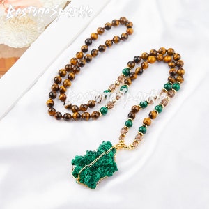 May include: A long necklace made of brown tiger's eye beads and green malachite beads with a large green malachite pendant. The necklace is on a white fabric background.
