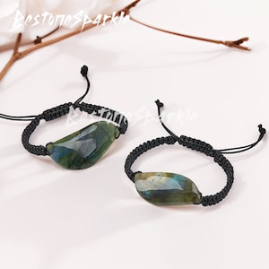 May include: Two black braided bracelets with a green and blue labradorite stone pendant. The bracelets are adjustable with a sliding knot.
