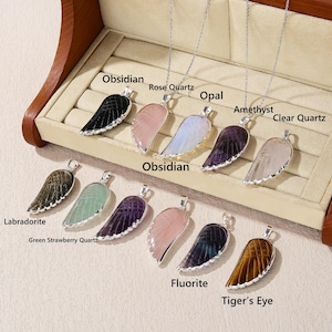 May include: A collection of gemstone wing pendants on silver chains. The pendants are in various colors, including black obsidian, rose quartz, opal, amethyst, and clear quartz. The image also includes Labradorite, Green Strawberry Quartz, Fluorite, and Tiger's Eye pendants.