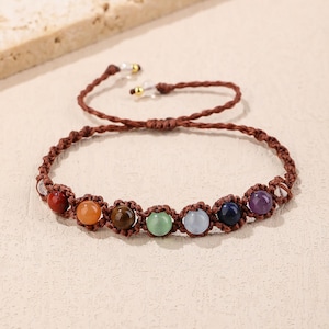 Könnte beinhalten: Braunes Makramee-Armband mit sieben farbigen Edelsteinperlen. Die Perlen sind in einer Reihe angeordnet und sind rot, orange, braun, grün, hellblau, dunkelblau und lila. Verstellbarer Knotenverschluss.