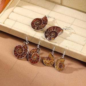 May include: A collection of fossilized ammonite shell earrings. The earrings feature a spiral design with shades of brown and tan, set in silver-colored metal. The earrings are displayed in a beige jewelry box.