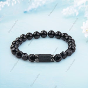 Op de afbeelding: Zwarte kralen armband met een zwarte steen accent en een zilveren metalen kraal met zwarte stenen. De armband ligt op een lichtblauwe achtergrond.