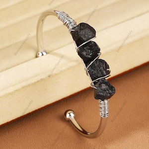 Puede incluir: Una pulsera de puño plateada con tres piedras negras en bruto sujetas con alambre plateado. La pulsera tiene un diseño curvo con un extremo redondeado y una bola decorativa. La pulsera se muestra sobre una superficie marrón claro.