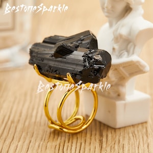 May include: A black tourmaline crystal ring with a gold band. The ring is made of two pieces of tourmaline that are connected by a gold band. The ring is on a wooden surface.