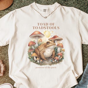 Könnte beinhalten: Ein elfenbeinfarbenes Kurzarm-T-Shirt mit einem Grafikdruck eines braunen Kröte, umgeben von roten und weißen Pilzen, einer gelben Sonne und grünem Laub. Der Text lautet "TOAD OF TOADSTOOLS" über der Grafik und "protector of the patch" darunter.
