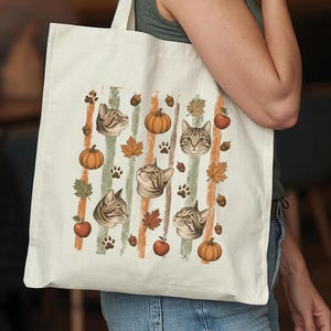 May include: A cream-colored canvas tote bag featuring a repeating pattern of brown tabby cat faces, orange pumpkins, red apples, brown acorns, maple leaves, and paw prints arranged in vertical stripes against a light background.