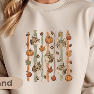May include: A sand-colored crewneck sweatshirt featuring a central graphic print of tabby cat faces, orange pumpkins, red apples, brown acorns, autumn leaves, and paw prints arranged in vertical stripes against a light background.