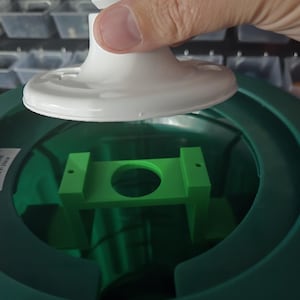 May include: A white light fixture with a round base is being installed into a green plastic structure. The fixture has an ETL Intertek label. A green plastic bracket is visible inside the structure.