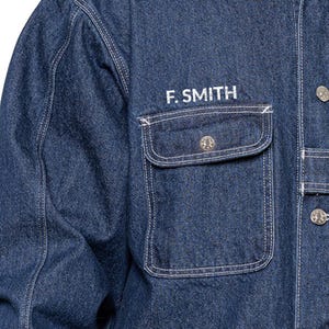 May include: Close-up of a denim jacket with the embroidered text "F. SMITH" above a pocket. The jacket is a medium blue color with white stitching and silver buttons. The pocket has a decorative button.