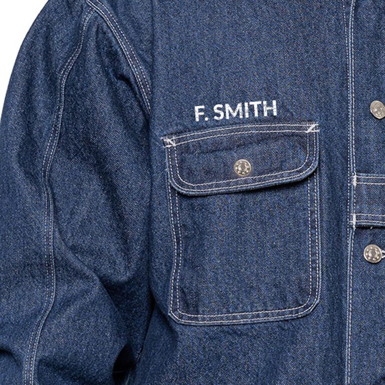 May include: Close-up of a denim jacket with the embroidered text "F. SMITH" above a pocket. The jacket is a medium blue color with white stitching and silver buttons. The pocket has a decorative button.