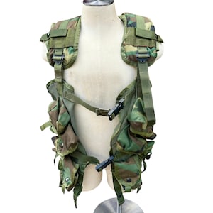 May include: Camouflage tactical vest with multiple pockets and adjustable straps. The vest features a woodland camouflage pattern in shades of green, brown, and tan. It has black plastic buckles and is displayed on a mannequin.
