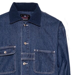 May include: A dark blue denim jacket with a contrasting navy blue corduroy collar. The jacket features two chest pockets and two lower pockets, all with white stitching. The jacket has silver buttons and the brand name "GAME" is visible on a tag.