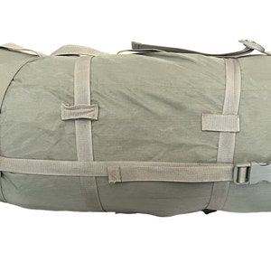 May include: A cylindrical, olive-green duffel bag with tan straps and a side-release buckle. The bag has two top handles and two straps that wrap around the circumference. The bag is made of a lightweight, possibly water-resistant material.