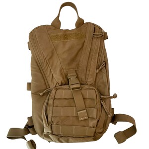 Op de afbeelding: Een bruinbeige rugzak met meerdere compartimenten en riemen. Voorzien van een handvat aan de bovenkant, een voorvak met ritssluiting en verstelbare schouderbanden. De rugzak heeft een tactisch ontwerp met een gespsluiting en een klittenband patch.
