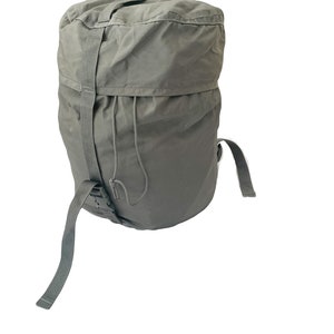 Könnte beinhalten: Ein grauer Rucksack mit Kordelzugverschluss und verstellbaren Trägern. Der Rucksack hat eine obere Klappe und einen einzelnen Riemen, der über die Vorderseite verläuft. Die Träger sind aus strapazierfähigem Stoff.