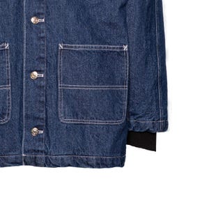 May include: Close-up of a dark blue denim jacket with silver buttons and white stitching. The jacket has two large front pockets and a black ribbed cuff on the sleeve. The fabric has a textured appearance.