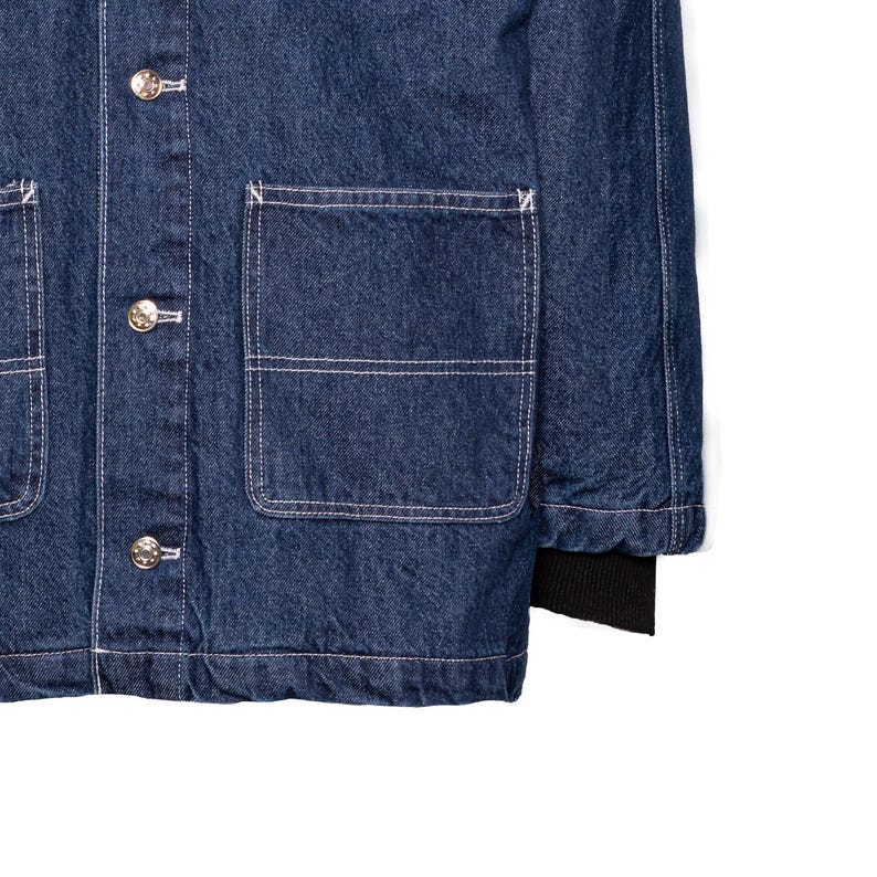 May include: Close-up of a dark blue denim jacket with silver buttons and white stitching. The jacket has two large front pockets and a black ribbed cuff on the sleeve. The fabric has a textured appearance.