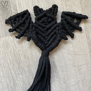 Könnte beinhalten: Schwarzer Makramee-Fledermaus-Wandbehang mit einer langen Quaste.