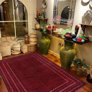 May include: A red and black patterned rug with a detailed border. The rug is laid out on a wooden floor in a room with a decorative mirror and a shelf with various vases and other decorative items.