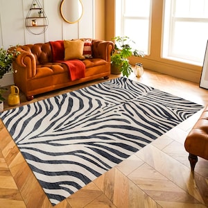 May include: A black and white zebra print area rug with a cream background. The rug is laid out on a hardwood floor in a living room setting with a brown leather sofa and plants.