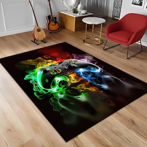 Puede incluir: Una alfombra rectangular negra con una imagen estilizada de un mando de videojuegos. El mando es negro con detalles dorados, rodeado de humo colorido y arremolinado en tonos verdes, amarillos, azules y rojos. La alfombra está sobre un suelo de madera.