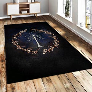 May include: A rectangular black rug featuring a white tree design with a circular inscription in a gold color. The tree is centered on a dark blue background, with the inscription surrounding it. The rug is placed on a wooden floor.