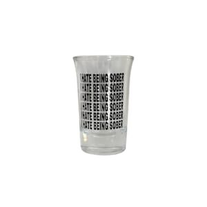 May include: A clear shot glass with the phrase "I HATE BEING SOBER" printed in black, repeated multiple times. The glass has a slightly flared shape.