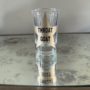 May include: A clear glass shot glass with a gold star sticker that says "Throat Goat" on it. The shot glass is sitting on a table with a reflective surface.